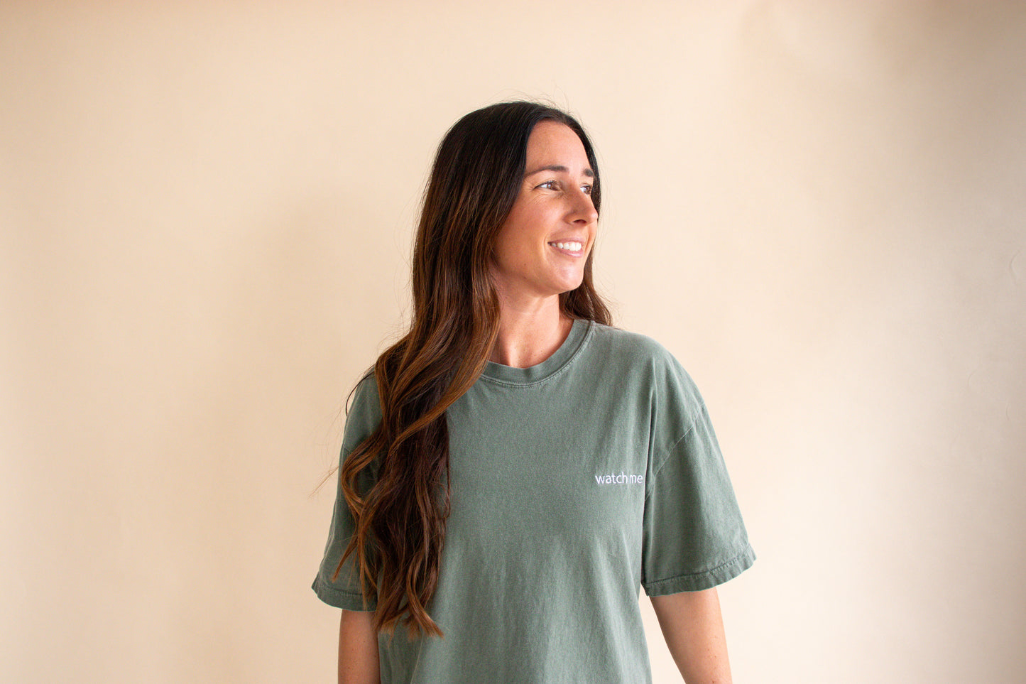 Woman wearing a green t-shirt with the words watch me on left chest on a beige background