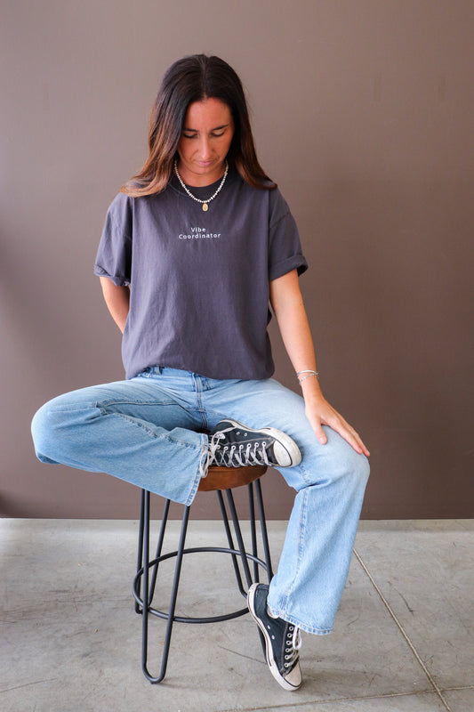 Woman sitting on a stool wearing a dark gray t-shirt with embroidery saying vibe coordinator and light blue jeans.