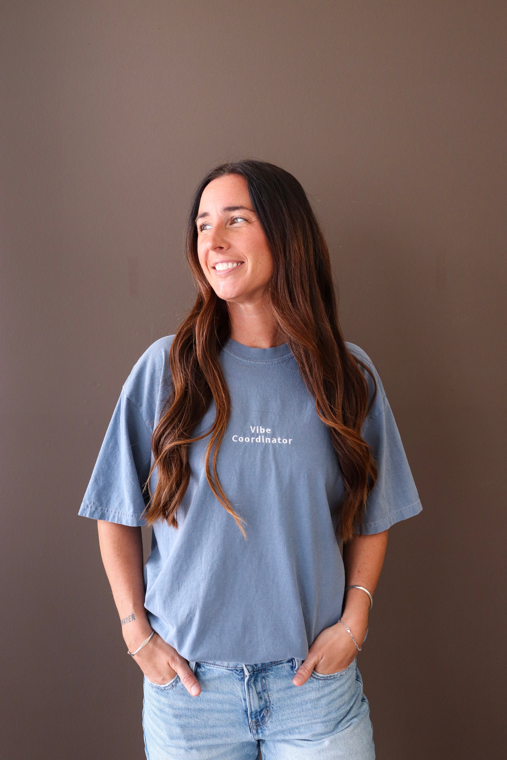 Woman wearing a blue t-shirt with text saying vibe coordinator, standing against a brown background