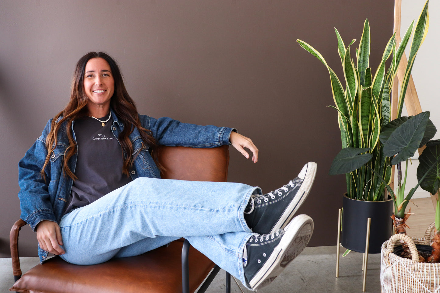 Woman sitting in a chair with plants in the background wearing grey shirt that says vibe coordinator