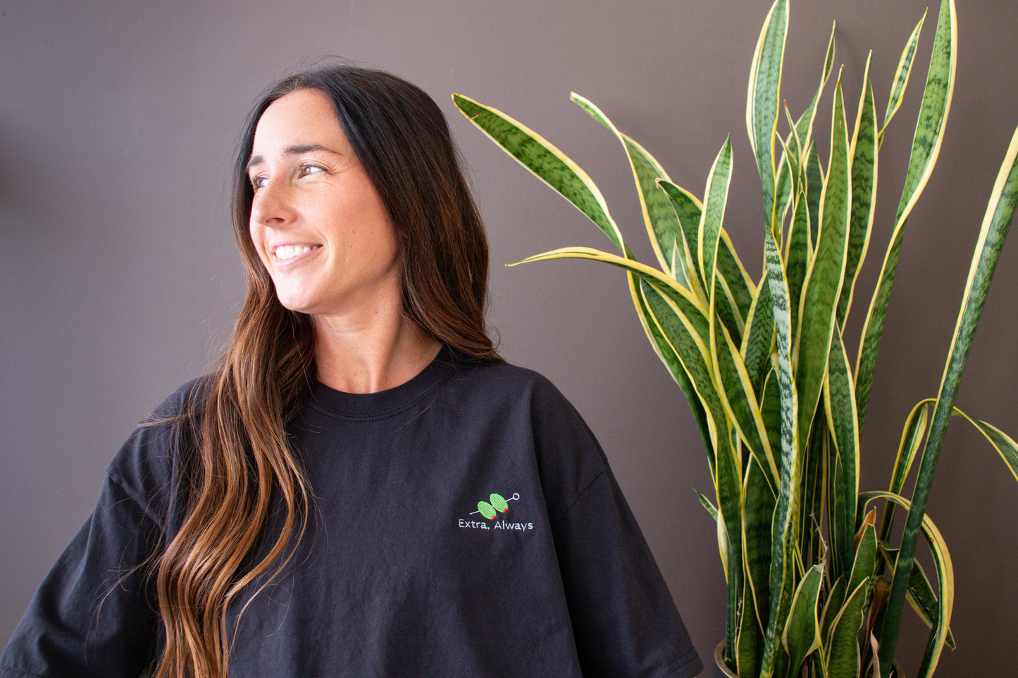 Woman wearing an Extra, Always black shirt with a logo next to a potted plant against a gray background