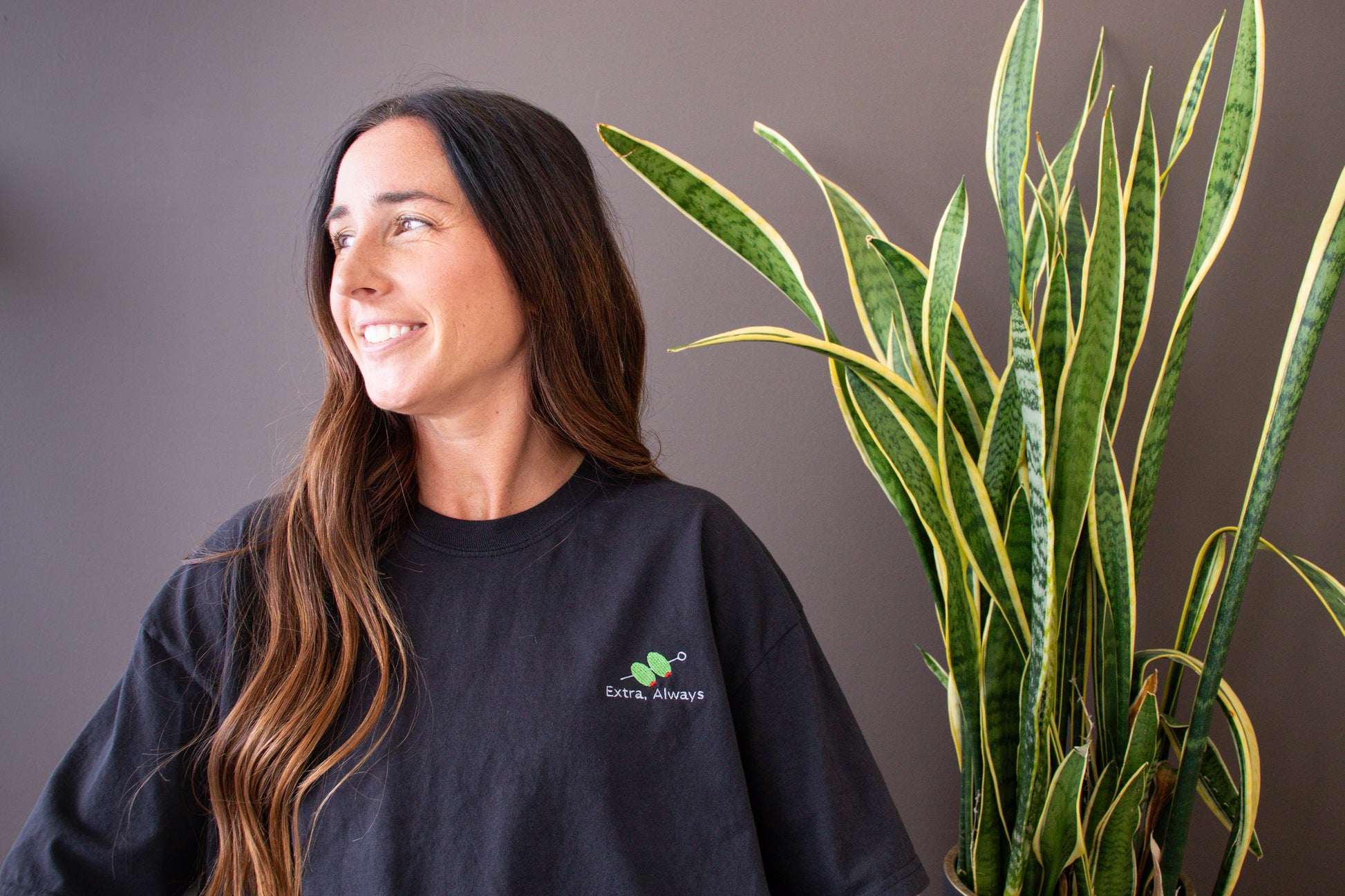 Woman wearing an Extra, Always black shirt with a logo next to a potted plant against a gray background