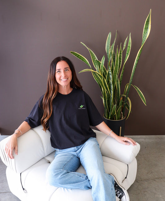 Woman sitting on white couch with brown wall wearing Extra, Always t-shirt