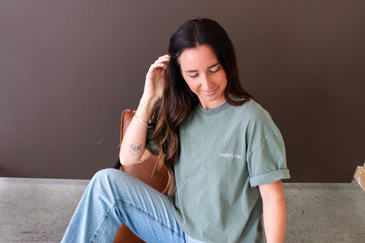 Woman wearing a green t-shirt with text saying watch me on left chest, sitting on a concrete surface against a brown wall.